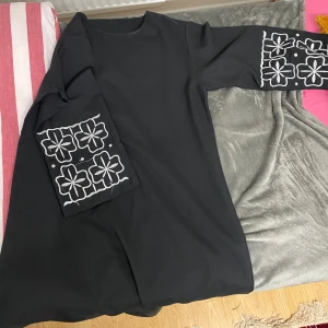 Svart abaya med blommigt mönster - Elegant svart abaya med lång ärm och vitt blommigt mönster på ärmarna. Perfekt för en stilren look.