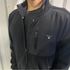 GANT fleecejacka  - Snygg svart fleecejacka från GANT med dragkedja och hög krage. Jackan har en praktisk bröstficka med dragkedja och broderad logotyp på bröstet. Perfekt för kyliga dagar.