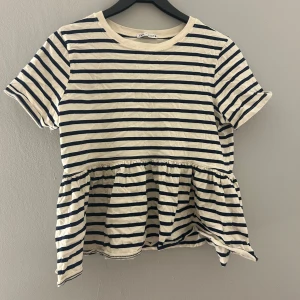 Randig topp från Zara - Säljer en söt randig topp från Zara i beige och blått. Toppen har korta ärmar och en volangdetalj vid midjan som ger en fin form. Perfekt för en avslappnad stil.