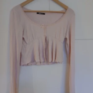 Beige långärmad topp från Gina Tricot - Säljer en söt beige långärmad topp från Gina Tricot. Toppen har en liten rosett framtill och är croppad med volangdetaljer. Perfekt för en stilren och feminin look.