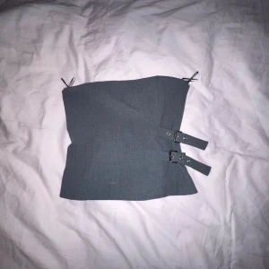 Grå korsett med spännen - Snygg grå korsett med två dekorativa spännen på sidan. Perfekt för att ge en edgy touch till din outfit. Den har en stilren design och smala axelband.