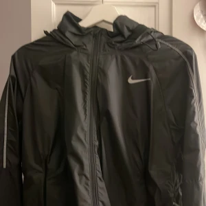 Svart vindjacka från Nike - Snygg svart vindjacka från Nike med huva och dragkedja. Perfekt för blåsiga dagar. Jackan har en stilren design med Nike-logga på bröstet.