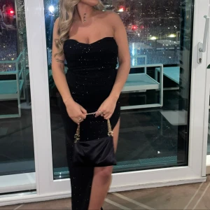 Svart glittrig axelbandslös klänning - Snygg svart axelbandslös klänning med glittrande detaljer. Klänningen har en slits på sidan som ger en elegant touch. Perfekt för festliga tillfällen.