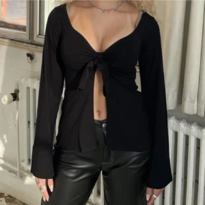 Svart långärmad topp från Design by Si - Snygg svart långärmad topp från Design by Si med knytning framtill. Perfekt för en stilren look med en touch av elegans. Toppen har en v-ringad design och är tillverkad i ett mjukt material.
