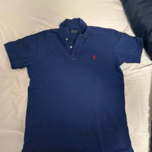 Blå pikétröja från Ralph Lauren - Säljer en klassisk blå pikétröja från Ralph Lauren med röd broderad logga på bröstet. Tröjan har korta ärmar och knappar vid kragen. Perfekt för en stilren look.