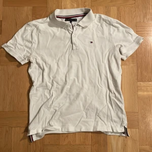 Vit pikétröja från Tommy Hilfiger - Snygg vit pikétröja från Tommy Hilfiger med klassisk krage och knappslå. Den har en liten logotyp på bröstet och ränder i kragen. 