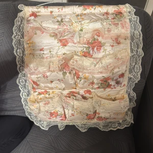 Blommig förvaringsväska med spetsdetaljer - Charmig förvaringsväska i blommigt mönster med spetsdetaljer. Perfekt för att organisera småsaker. Väskan har flera fickor och en dekorativ rosett.