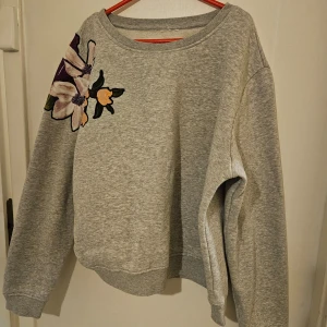 Grå sweatshirt med blommigt tryck - Säljer en grå sweatshirt med ett stort blommigt tryck på ena axeln. Tröjan har långa ärmar och är tillverkad i en blandning av bomull och polyester. Perfekt för en avslappnad stil.