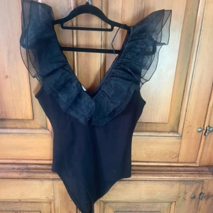 Svart body med volangdetaljer - Snygg svart body med djup v-ringning och volangdetaljer i transparent tyg vid axlarna. Perfekt för en stilren och elegant look. (Storlek L men sitter mer som en s/m)