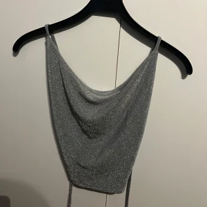 Glittrig silverfärgad linnetopp - Snygg glittrig silverfärgad linnetopp med tunna axelband och draperad framsida. Toppen har en öppen rygg med snörning för en justerbar passform. Perfekt för en festlig kväll! Svårt att ta bild bakifrån men sitter så snyggt fram och korsar där bak. Fin att ha till fester osv. 