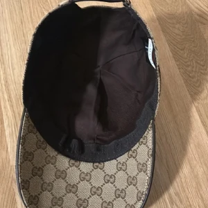 Beige keps från Gucci - Snygg beige keps från Gucci med det klassiska GG-mönstret. Kepsen har en justerbar rem baktill och en röd och grön randdetalj på sidan. Perfekt för att ge en lyxig touch till din outfit., Får med låda Å kvitto!!