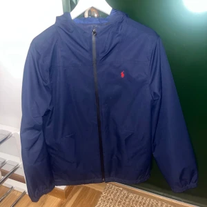 Mörkblå jacka från Polo Ralph Lauren - Mörkblå vind/regnjacka från polo Ralph lauren med röd logga på bröstet. Den är i barnstorlek XL men är förstor för mig som är tjej med storlek S. Använd fåtal gånger så är i mycket bra skick. 