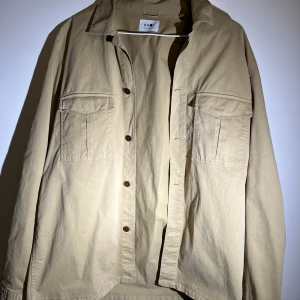Beige overshirt från NN07 - Snygg beige overshirt från NN07 med klassisk krage och knappar framtill. Två praktiska bröstfickor ger en stilren look. Perfekt för lager-på-lager-stil.