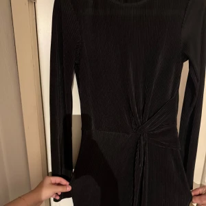 Svart ribbad festklänning - Elegant svart ribbad festklänning med långärmad design och knytning i midjan från Nelly. Lång och delar sig framifrån. Perfekt för en stilren look. Endast använd vid två tillfällen.