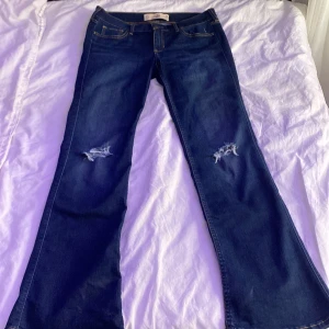 Mörkblå low-waisted bootcut jeans från Hollister - Snygga mörkblå bootcut jeans från Hollister med slitna detaljer på knäna. Perfekta för en avslappnad stil. Klassisk femficksdesign och knappgylf.