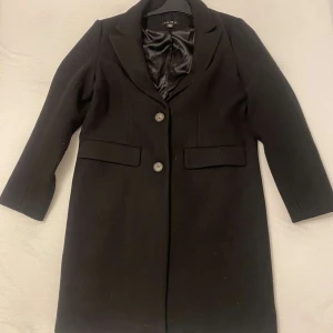 Svart kappa från Amisu - Elegant svart kappa från Amisu med knappar framtill och klassisk krage. Perfekt för en stilren look. Kappan har långa ärmar och är tillverkad i ett mjukt material.