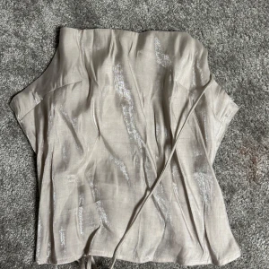 Beige glittrig tubtopp - Snygg beige tubtopp med glittriga detaljer. Perfekt för en kväll ute eller festliga tillfällen. Toppen har en enkel design med en smickrande passform.