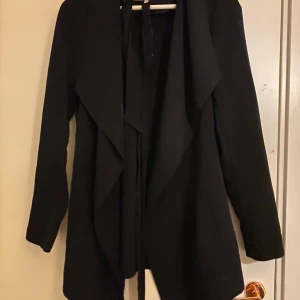 Svart kappa från VILA - Snygg svart kappa från VILA med knytband framtill. Perfekt för höst och vår med sin stilrena design och långa ärmar. En mångsidig jacka som passar till många olika outfits.