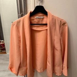 Persikofärgad kavaj från Soul River - Snygg tunn persikofärgad kavaj från Soul River med trekvartsärmar. Perfekt för en stilren look. Kavajen har en enkel design utan knappar och är tillverkad i ett lätt material.