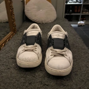 Valentino sneakers med mörkblå detalj - Snygga Valentino sneakers i vitt med en mörkblå  rem över snörningen. Skorna har en klassisk design med rund tå och platt sula. Perfekta inför sommarn. Dom är lite använda därav priset . Pris kan diskuteras vid snabb affär!