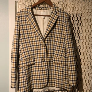 Rutig kavaj från H&M - Snygg rutig kavaj från H&M i beige, blå och vit. Kavajen har en klassisk krage och knäpps med knappar framtill. Perfekt för en stilren look.