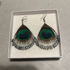 Pampiga påfågelfjäder-örhängen - Säljer ett par unika örhängen med påfågelfjädrar. De har en fantastisk färgkombination av grönt, blått och brunt, och är dekorerade med små pärlor längs kanterna. Perfekta för att ge en färgklick till din outfit! 🦚