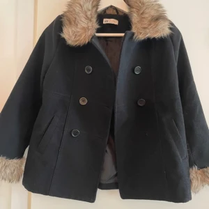 Svart kappa med fuskpäls från H&M - Säljer en snygg svart kappa från H&M i storlek 140 (9-10 år). Den har en mysig fuskpäls på kragen och ärmsluten, perfekt för kyliga dagar. Kappan är dubbelknäppt med stora knappar och har en klassisk stil. Passar perfekt till både vardag och fest! Har tappet en knapp längst ner men det är inget man märker eller som påverkats användningen. 