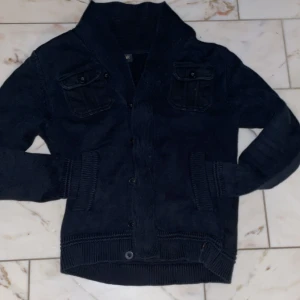 Svart kofta med knappar - Säljer en snygg svart cardiganen med knappar framtill och två bröstfickor med knappdetaljer. Den har en ribbad krage och muddar, vilket ger en stilren look. Perfekt för höst och vinter! 