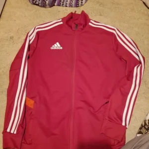 Röd träningsjacka från Adidas - Säljer en snygg röd träningsjacka från Adidas med klassiska vita ränder längs ärmarna. Jackan har dragkedja och är perfekt för träning eller vardagsbruk. Den är i mycket bra skick och har en normal passform. Perfekt för höst och vår!