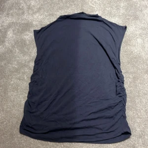 Mörkblå topp från Gina Tricot - Säljer en stilren mörkblå topp från Gina Tricot i storlek L. Toppen har en enkel design med rynkade detaljer på sidorna som ger en smickrande passform och lite högre nacke . Perfekt för både vardag och festliga tillfällen! Aldrig använd!
