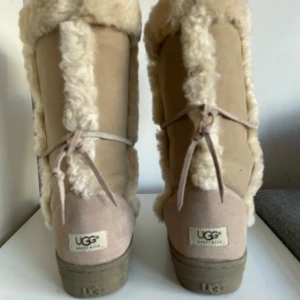 Beige UGG boots med päls - Säljer ett par mysiga höga UGG boots i beige mocka med fluffig pälsdetalj. Perfekta för kalla vinterdagar! De har en bekväm passform och är i mycket bra skick. Snörning baktill för extra stil och komfort.