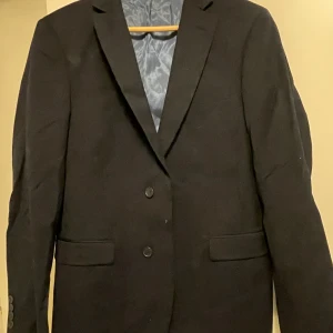 Svart kavaj från Dressmann - Säljer en stilren svart kavaj från Dressmann i storlek 48 Short. Kavajen har två knappar framtill och är perfekt för både formella och semi-formella tillfällen. Den är i mycket bra skick och passar utmärkt för höst och vår. Perfekt för fest eller bal!