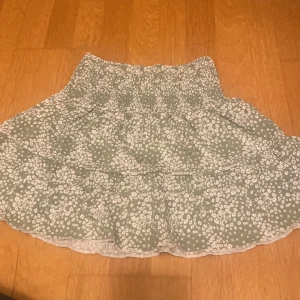 Blommig volang skjol - Söt kortkjol i ljusgrön med vita blommor. Kjolen har en volangdetalj och är perfekt för sommardagar. Tillverkad i mjuk bomull för en bekväm passform. Perfekt för en avslappnad dag på stan eller en picknick i parken.