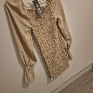 Beige klänning från Boohoo - Säljer en superfin beige klänning från Boohoo i nytt skick. Den har långa ärmar med smockdetaljer vid ärmslut och en tight passform. Perfekt för höst och vår! Klänningen är gjord i ett mjukt material som känns skönt mot huden.