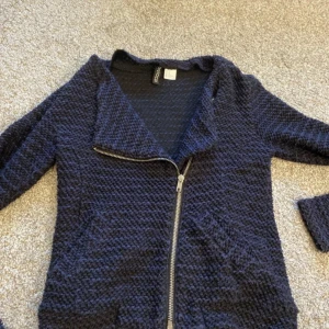 Mörkblå stickad kofta med dragkedja - Säljer en snygg mörkblå stickad jacka med asymmetrisk dragkedja. Jackan har en texturerad yta och är perfekt för höst och vår. Den är gjord av en blandning av polyester och bomull, vilket gör den både bekväm och hållbar. Perfekt för en casual look!