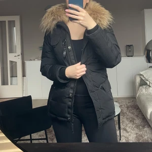 Svart dunjacka med pälsdetalj - Säljer en snygg svart dunjacka från Hollies med en fluffig pälsdetalj på huvan i bra skick, storlek 38. Jackan har långa ärmar och praktiska fickor med knappar. Bekväm och varm, med bra passform.  Använd, men i gott skick utan några synliga skador eller defekter.