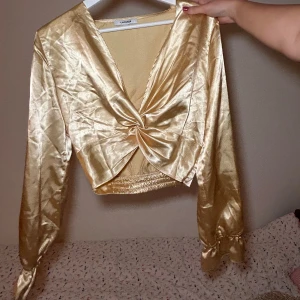 Guldig omlottopp från Chiquelle - Säljer en superfin guldig omlottopp från Chiquelle. Den har en glansig finish och långa ärmar med knytning vid ärmsluten. Perfekt för festliga tillfällen! ✨