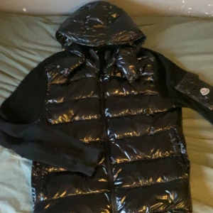 Svart dunjacka från Moncler - Säljer en snygg svart dunjacka från Moncler. Den har en glansig finish och är perfekt för kalla dagar. Jackan har en huva och långa ärmar med Moncler-logga på ärmen. Den är i mycket bra skick och passar perfekt för både höst och vinter.