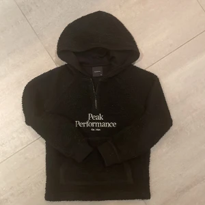 Svart teddy hoodie från Peak Performance - Säljer en svart Teddy hoodie från Peak Performance i fint skick. Den är mjuk och har en dragkedja vid halsen. Perfekt för kyliga dagar och har en stor, mysig huva. Passar både till vardags och för en avslappnad stil.