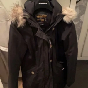 Svart parkajacka från Woolrich - Säljer en svart parkajacka från Woolrich i mycket bra skick. Jackan har en mysig pälsfodrad huva och stängs med både dragkedja och knappar. Perfekt för kalla vinterdagar! 🖤