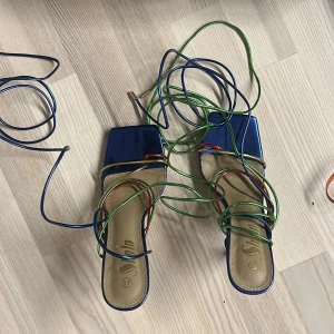 Färgglada klackskor med snörning - Snygga klackskor i blått med färgglada snören i grönt, blått och orange. De har en spetsig tå och hög klack, perfekt för festliga tillfällen. Skorna är i mycket bra skick och ger en unik touch till din outfit.