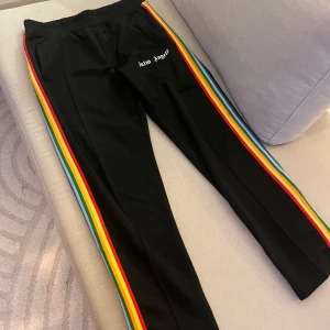 Svarta byxor med regnbågsränder från Palm Angels - Säljer ett par svarta byxor från Palm Angels med färgglada regnbågsränder längs sidorna. Byxorna har en elastisk midja och dragkedjor vid bensluten för extra stil. Perfekta för en avslappnad look med en touch av färg. 🖤🌈