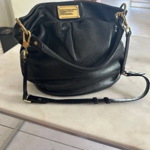 Svart axelväska i skinn - Säljer en snygg svart axelväska i skinn med gulddetaljer. Väskan har en justerbar axelrem och en platta med logotyp framtill. Perfekt för både vardag och fest! 🖤