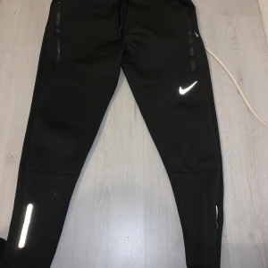 Svarta träningsbyxor från Nike - Säljer ett par svarta träningsbyxor från Nike i mycket bra skick. De har en tight passform och är perfekta för träning. Byxorna har reflexdetaljer och en dragkedja vid bensluten för extra komfort. Perfekta för höst- och vinterträning!