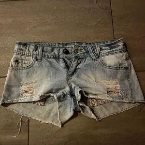 Lågmidjade jeansshorts - Säljer ett par ljusa lågmidjade jeansshorts med coola slitningar och råa kanter. De har en avslappnad look med blommigt tyg som syns genom slitningarna. Perfekta för sommardagar! 🩷🫶🏼. Midjemåttet tvärs över är 39cm