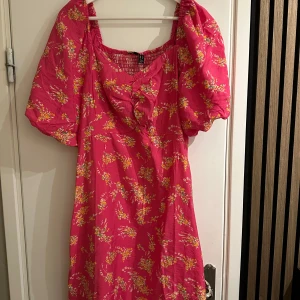 Rosa blommig klänning från Vero Moda - Superfin rosa klänning från Vero Moda med blommigt mönster i gult och vitt. Helt oanvänd, endast testad. Den har puffiga kortärmar och en smockad rygg för en bekväm passform. Perfekt för sommarens fester eller en dag på stan. Tillverkad i 100% polyester.
