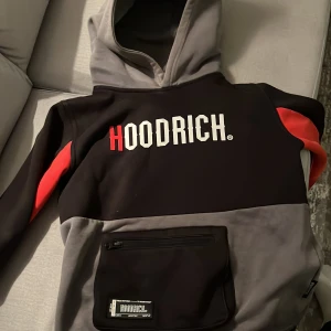 Svart och grå hoodie från Hoodrich - Säljer en snygg svart och grå hoodie från Hoodrich med röd text och detaljer. Den har en stor ficka framtill och en bekväm huva. Perfekt för en casual look eller träning. Passar bra till höst och vinter.