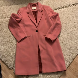 Rosa kappa från Gant - Säljer en superfin rosa kappa från Gant i mycket bra skick. Den har en klassisk design med en enkel knapp framtill och långa ärmar. Passar perfekt till både vardag och fest!