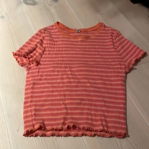 Randig topp från Uniqlo - Säljer en superfin randig topp från Uniqlo i orange och rosa. Den har korta ärmar och en söt volangdetalj vid ärmslut och nederkant. Perfekt för sommardagar eller en avslappnad vårlook. Toppen är i bra skick och redo för en ny ägare!