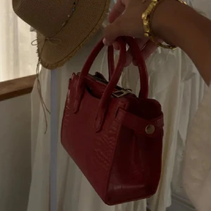 Väska i Burgundy  - Säljer en snygg röd handväska i skinn. Den har en stilren design med två handtag och en dragkedja upptill. Perfekt för att ge en färgklick till din outfit! 👜